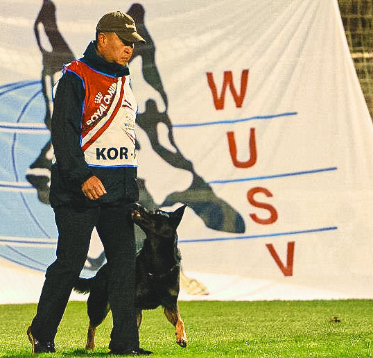 Ki Hong, wearing a team jacket emblazoned with 'KOR' and a baseball cap, confidently strides across the field with a black German Shepherd by his side at the WUSV World Championship. Behind them, a large banner with the letters 'WUSV' serves as a powerful backdrop, signifying the global scale of the competition.