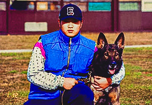 Steve Hong, as a young adult, wearing a blue and white jacket and a baseball cap, is kneeling on a grassy field beside his alert German Shepherd. Both are posing for the camera, with the dog's ears perked up and Steve's, engaging expression.