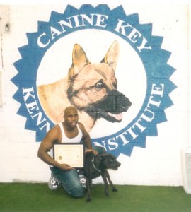 Eddie, proudly kneeling beside a graduated German Shepherd in front of the Canine Key Institute emblem, holding a certificate of completion