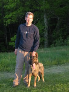 A person stands proudly in an open field during sunset with Kara, a German Shepherd, at their side. Both are looking towards the camera, with the dog's tongue playfully out, suggesting a moment of relaxation after training.